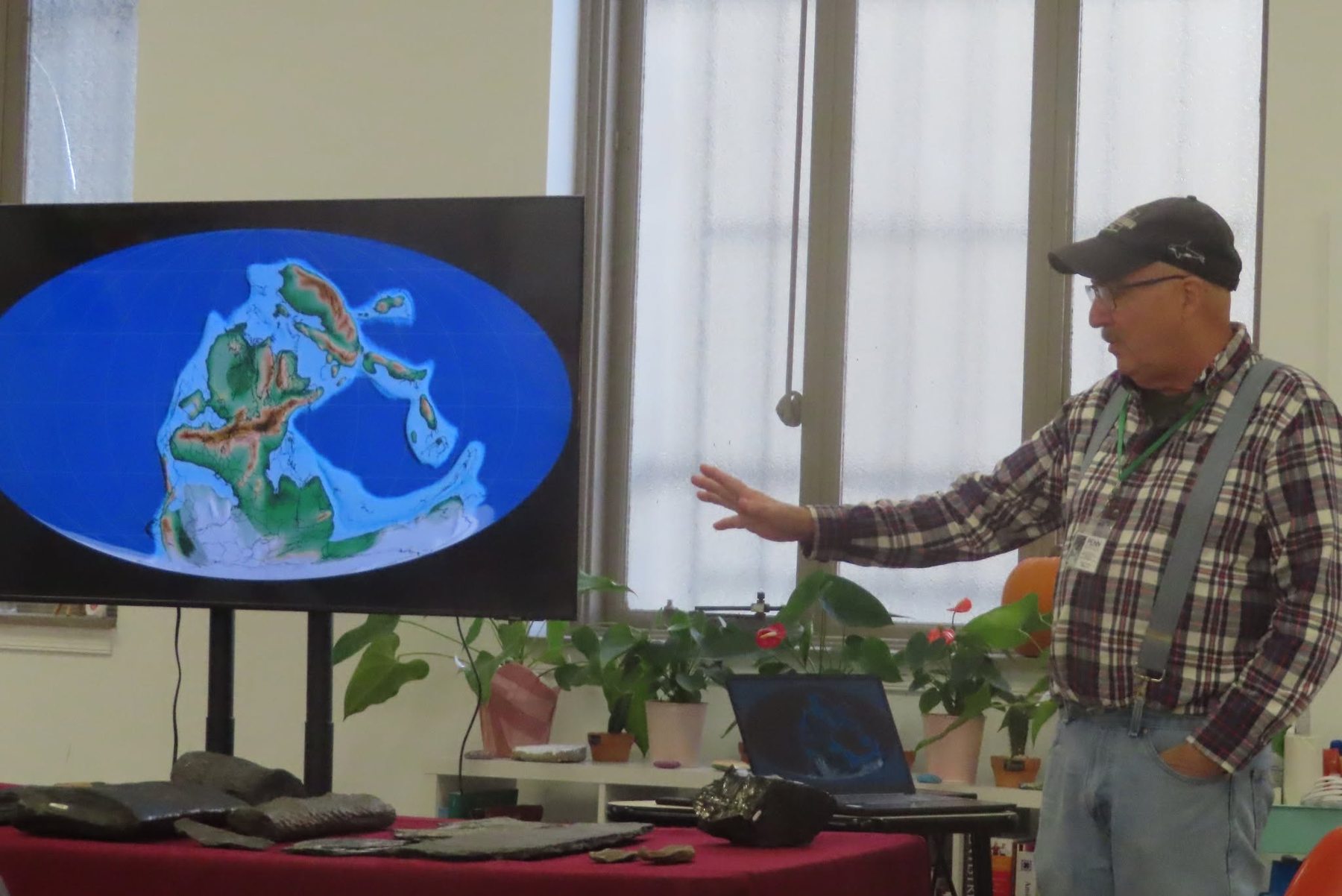 A member giving a talk on Pangea and Pennsylvania geology, gesturing at a continental map on screen with fossils on the table